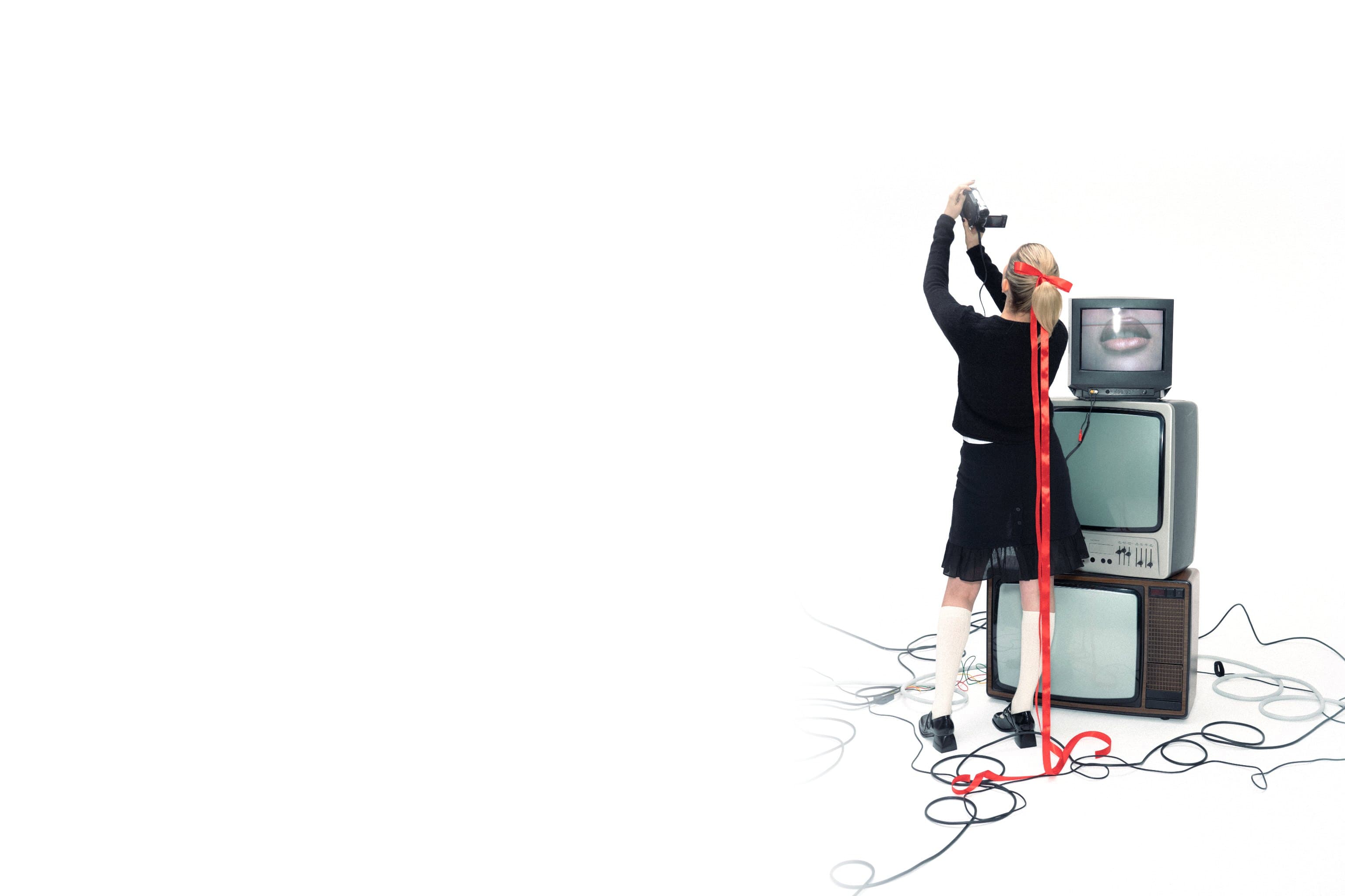 A person in black filming a stack of vintage televisions, a red ribbon trailing through a tangle of cables